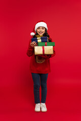 Wall Mural - full length of happy girl in santa hat and sweater holding bunch of Christmas presents on red