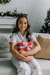 Wall Mural - Happy kid in dotted pajama holding New Year gift and sitting on couch