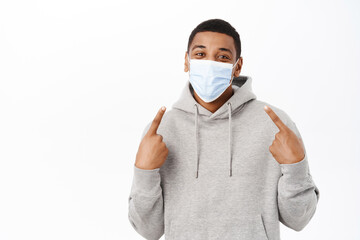 Wall Mural - Covid awareness, healthcare concept. Image of African american man put on medical face mask to prevent coronavirus spread, standing over white background