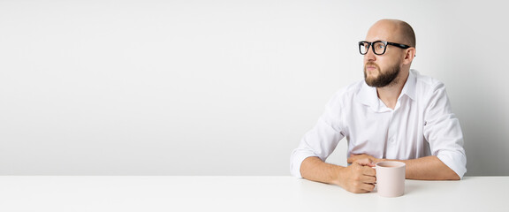 Pensive young man with a cup sitting at the table on a white background. Banner