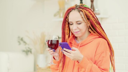 Wall Mural - Young woman with bright long dreadlocks browsing mobile phone, drinking red wine, dancing in kitchen.