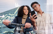 © N F/peopleimages.com - Travel, phone and city business people with overlay for digital, social media and data analysis of transportation urban report on eco scooter. Young carbon footprint worker or couple with seo review