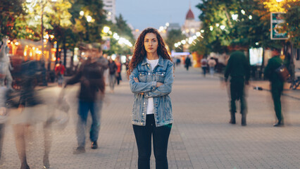 Portrait of stylish young lady tired of usual haste standing in the street among whizzing people and looking at camera. Time, youth and modern society concept.