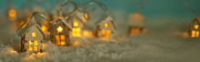 Abstract Christmas Winter Panorama With Wooden Houses Christmas String Lights In Cold Snow Landscape And Glowing Golden Lights In Background. Panorama, Banner