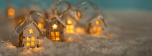 Abstract Christmas Winter Panorama With Wooden Houses Christmas String Lights In Cold Snow Landscape And Glowing Golden Lights In Background. Panorama, Banner. Christmas Or Energy Themes.