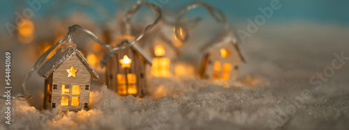 Fototapete Abstract Christmas Winter Panorama with Wooden Houses Christmas String Lights in Cold Snow Landscape and Glowing Golden Lights in Background. Panorama, Banner. Christmas or Energy themes.