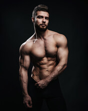 Slim Muscular Male Model At Black Background. Fitness Shirtless Guy In Black Sport Pants Posing In Studio. Man With Six Pack Abs Looking At Camera.