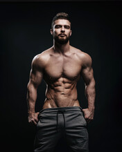 Slim Muscular Male Model At Black Background. Fitness Shirtless Guy In Black Sport Pants Posing In Studio. Man With Six Pack Abs Looking At Camera.