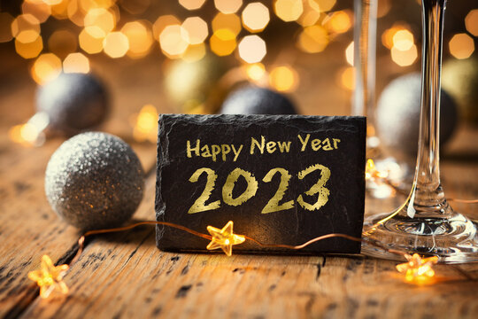 Fototapete - Happy New Year 2023 - Greeting Card - Black slate board with golden English text. Christmas decoration on rustic wood with Champagne glasses and golden  lights from fireworks in background