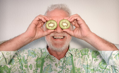 Wall Mural - Smiling bearded senior man covering his eyes with two half kiwi fruit. Funny portrait of senior caucasian male holding a kiwi cut in half over his eyes, white background