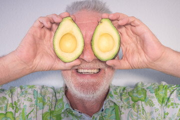 Wall Mural - Funny portrait of senior caucasian male holding an avocado cut in half over his eyes, white background. Smiling bearded senior man covering his eyes with two half apple fruit.