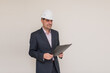 © KatrinaQQ - A male architect-builder in a strict suit and helmet with a plan and a pen in his hands looks at the camera, a male businessman on a gray background