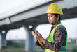 © anut21ng Stock - Asian construction worker wearing a helmet stands at construction site touching smartphone