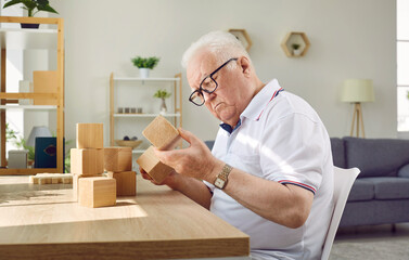 Patient with dementia playing game in geriatric clinic or nursing home. Senior man sitting at desk and looking at wooden cube that he is holding in hand. Old age, Alzheimer's disease, therapy concept