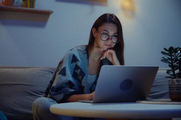 Young smiling woman using laptop at night, working overtime at home sitting on sofa, flexible working hours