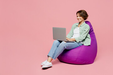 Full body young IT woman wear green shirt white t-shirt sit in bag chair hold use work on laptop pc computer isolated on plain pastel light pink background studio portrait. People lifestyle concept.