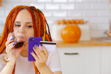 Wall Mural - Young woman with dreadlocks drinking red wine from glass, browsing mobile phone, entering card details, paying internet purchase, sitting in kitchen.