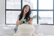 © kaew6566 - Cheerful asian woman playing ukulele sitting on white bed with pillows beside window at apartment. Joyful young girl holding acoustic ukulele in morning at home. Relax female play music ukulele hobby.