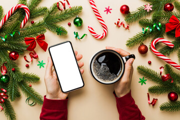 Wall Mural - Above view of female hand holding smart phone with hot cup of coffee. Christmas decoration on colored background