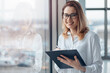 © mikelaptev - Young smiling woman standing at the panoramic window of a modern office and holding a folder with documents.