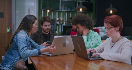 Friend showing laptop screen to work colleague seated at coffee shop. Group of diverse people using modern technological devices at cafe restaurant. Friends studying together working remotely