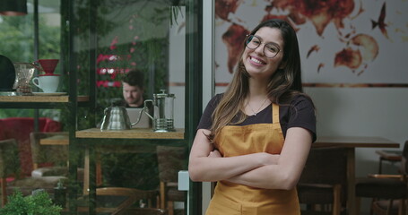 Happy female staff crossing arms smiling at camera in front of coffee shop store wearing yellow apron. A hispanic latin adult girl barista employee of small business restaurant place