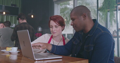 African American manager explaining job to new young female employee in front of laptop sitting at coffee shop. Two diverse staff collaborating together using computer. 