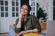 © Iona - Adorable African American girl in casual shirt sitting at desk with laptop looks at camera against blurry interior. Attractive Brazilian young woman remote working home. Business people and success.