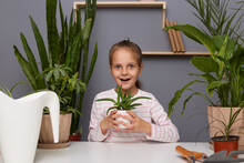 Indoor Shot Of Excited Amazed Little Cute Girl In Striped Shirt Sitting At Table Among Green Plants, Looking At Camera With Big Surprised Eyes, Holding Flower Pot With Favorite Plant.
