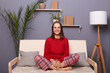 Indoor shot of happy delighted Caucasian woman wearing red sweater and checkered pants sitting on sofa with crossed legs in home interior, looking at camera with toothy smile.