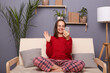 Portrait of attractive brown-haired woman blogger wearing red sweater and checkered pants sitting on cough in living room at home, holding mobile phone and waving hand, saying hello to followers.