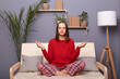 Portrait of calm young adult brown-haired woman wearing red sweater and checkered pants sitting on cough in lotus pose in living room at home, practicing yoga, relaxing.