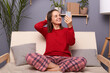 Portrait of smiling positive Caucasian woman wearing red sweater and checkered pants sitting on sofa in home interior, holding smart phone in hands, making selfie or having video call.