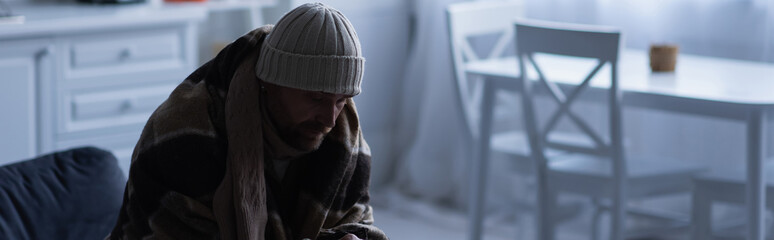 Wall Mural - frozen man sitting in warm blanket and knitted hat at home in twilight, banner.