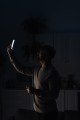 Wall Mural - man standing in dark kitchen with smartphone in raised hand and searching for connection during power blackout.