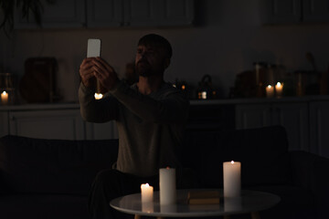 Wall Mural - man sitting near burning candles in dark kitchen and catching mobile connection on smartphone.