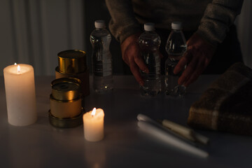 Wall Mural - cropped view of man holding bottled water near canned food and candles during energy blackout.