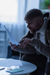 Wall Mural - frozen man in warm sweater and blanket looking at payment bills during electricity shutdown.