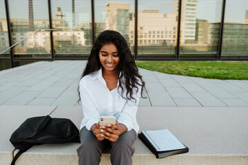 Wall Mural - Young indian woman using cellphone while sitting by building outdoors