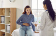 © Studio Romantic - Female patient with abdominal pain undergoing medical examination by doctor for indigestion. Young woman is talking to doctor and holding her stomach while sitting on medical couch in doctor's office.