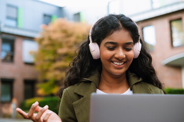 Wall Mural - Young woman using laptop and headphones while sitting on city street