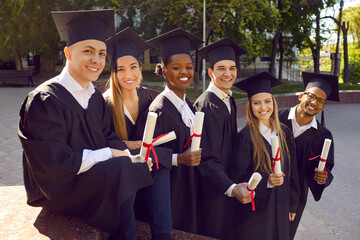 Wall Mural - Graduate.Young joyful people of different races and nationalities who have received diploma graduation from university or college are dressed in student black gown posing standing on street.