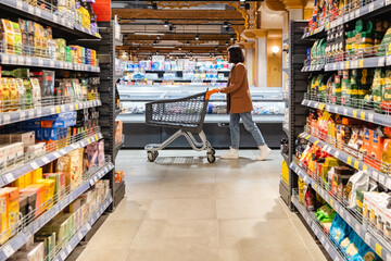 a woman with a cart walks between rows of shelves in a grocery store