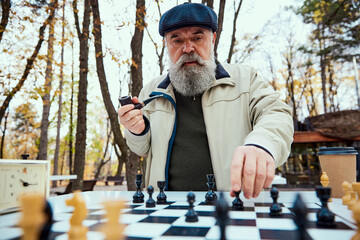 Wall Mural - Portrait of senior bearded man playing chess and smoking pipe in the park on a daytime in fall. Concept of leisure activity, old generation