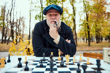 Wall Mural - Portrait of senior man playing chess in the park on a daytime in fall. Thinking and smoking pipe. Concept of leisure activity, old generation