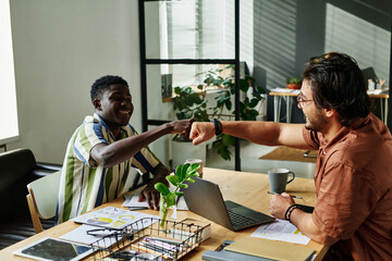 Wall Mural - Two happy young intercultural colleagues making punch bump over workplace while congratulating one another on successful deal