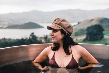 Wall Mural - blue-eyed caucasian young woman looking to the side wearing a swimsuit and a brown cap inside the small round jacuzzi with a view of the mountains and french farm bay, te wepu pods akaroa, new zealand