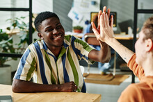 Young Successful African American Businessman Giving High Five To Female Colleague While Sitting By Workplace In Front Of Her