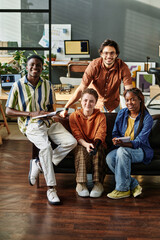 Team of young cheerful multicultural managers sitting on couch in office against their workplaces with computers and other supplies