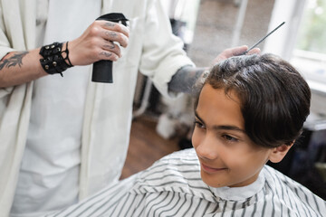 tattooed barber applying hair spray on smiling teenage client.